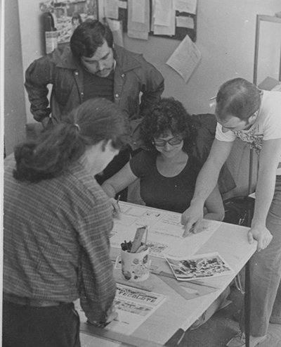 El Tecolote staff, including a young Juan Gonzales, stand over a drafting table and look at the newspaper